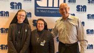 Two religious sister and man stand in front of SMLC logo