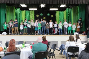 Row of children singing on stage with green backdrop