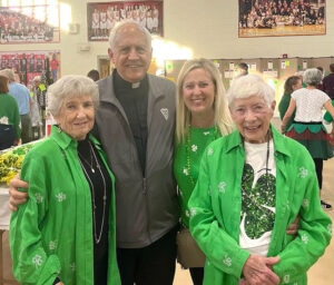 Three women in green and priest smiling at event in school gym